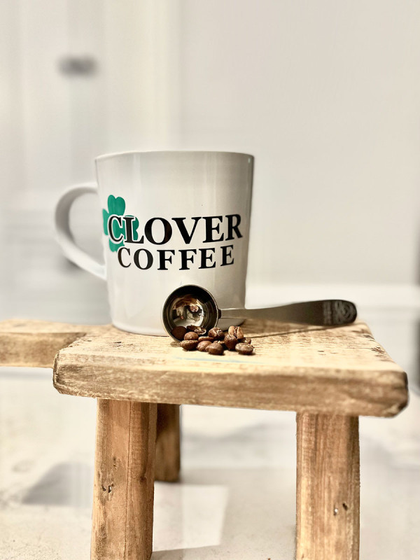 White mug with the text 'CLOVER COFFEE' and a green clover design, positioned on a rustic wooden stool. Beside the mug, a scoop with coffee beans is laid on the stool's surface, with a blurred white room background.